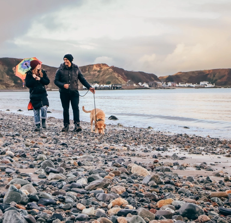 Couple and dog on a pebble beach beside a calm bay, with distant coastal village and hills under a cloudy sky.