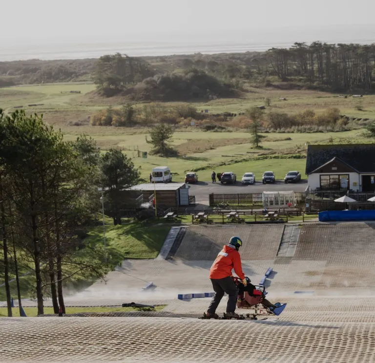 A child in an adapted sled / ski-pram being pushed by a man on a dry ski-slope.