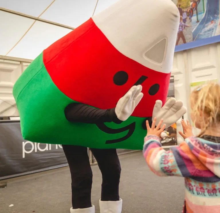 A costumed mascot in red and green leans down to greet a small child at an indoor event.