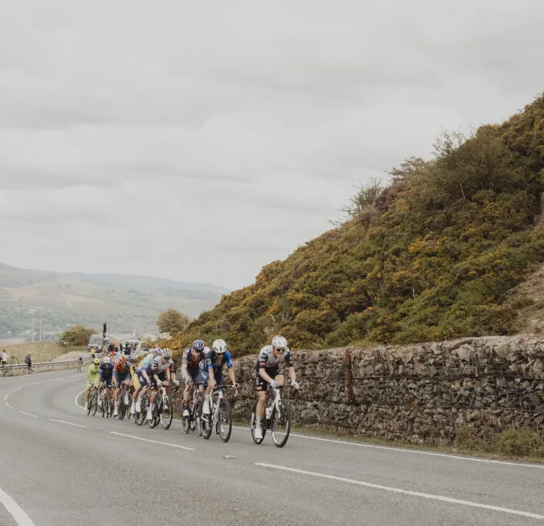 Professional cyclists racing in the 2025 Tour of Britain on the Tumble climb from Blaenavon, with a rocky hillside and gorse lining the road