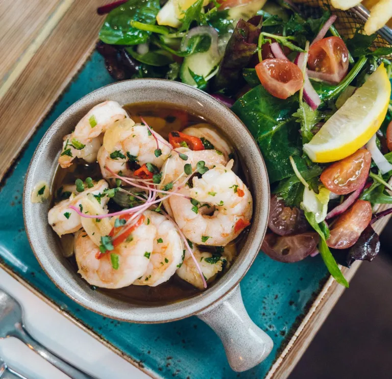 Bowl of prawns in a herb dressing served with mixed leaves, cherry tomatoes and a lemon wedge on a blue plate.