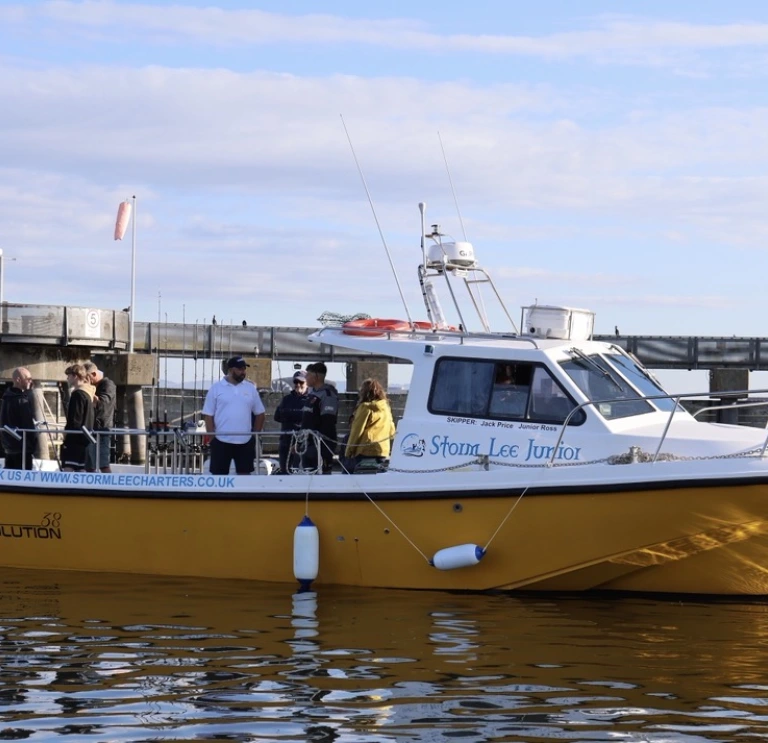 Passengers aboard the Storm Lee Junior charter boat on calm waters beside a harbour pontoon, with Cardiff Bay visible in the background under a partly cloudy sky.