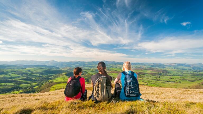 Three female walkers sat on the ground looking out across valleys and hills.