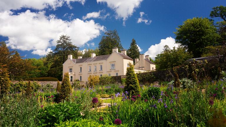 A cream coloured historic manor house surrounded by flower filled gardens