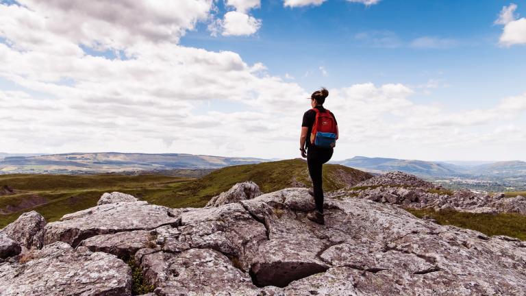 Wanderin auf einem Hügel stehend mit einem blauen Himmel im Hintergrund und mit Blick auf die grüne, bergige Landschaft der Brecon Beacons, Mittelwales