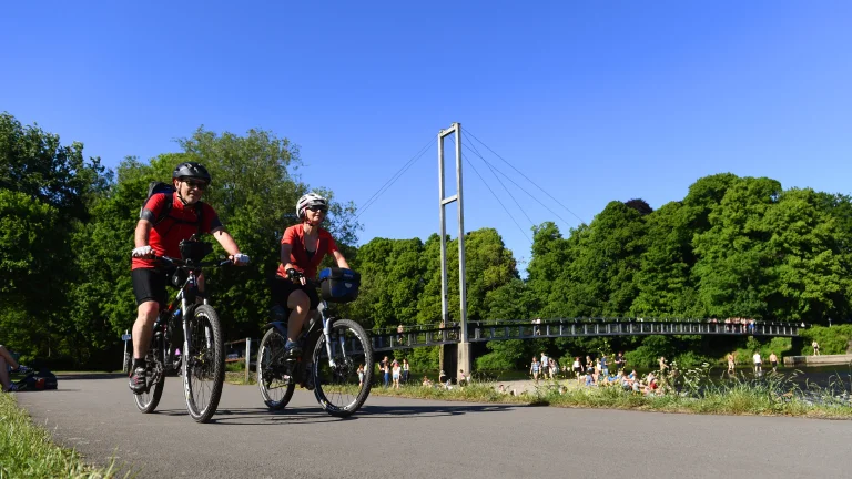 Two people cycling along a riverside path, with a modern footbridge and green trees beside the water under a clear blue sky.