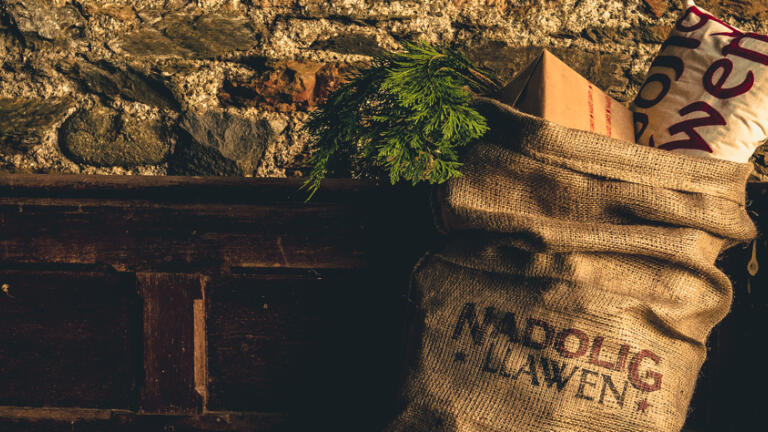 Brown hessian sack stuffed with christmas gifts and printed with the Welsh words Nadolig Llawen, hanging on a wooden mantle against a stone wall.