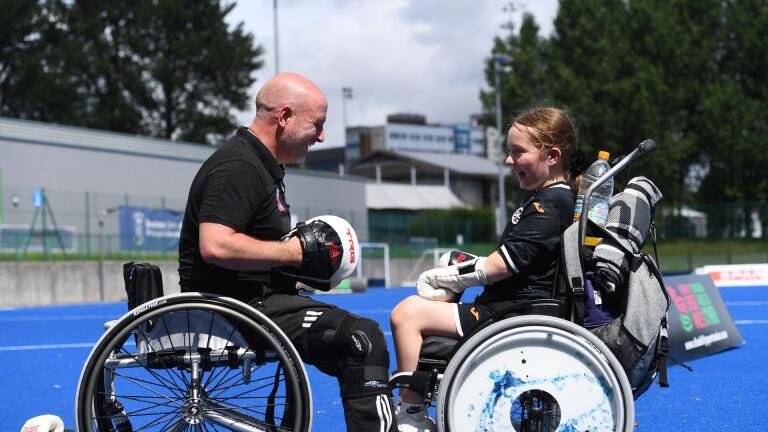 An man and a young girl in wheelchairs facing each other wearing boxing gloves and smiling.