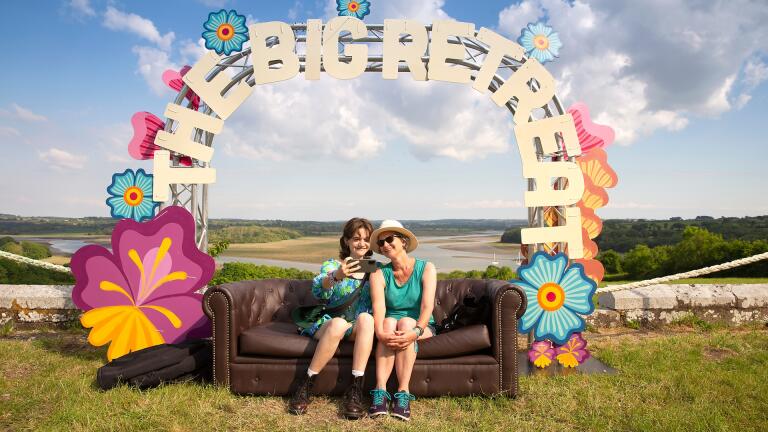 two woman sat on a sofa under a festival sign The Big Retreat, with views of the an estuary in the background.