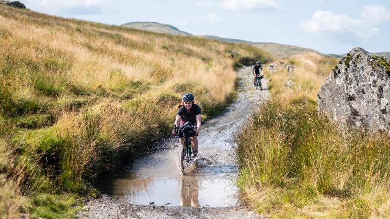 A gravel biker riding through a muddy puddle on a hillside track.