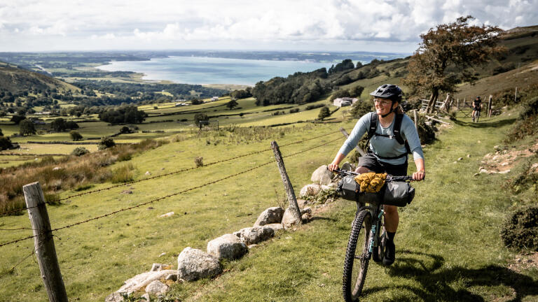 A female cyclist on a grassy roadway above a valley, looking down over the coast.