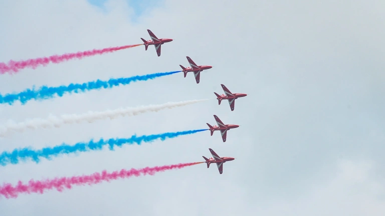 Five areoplanes, the Red Arrows, flying in an arrow formation with red white and blue vapour trails. 