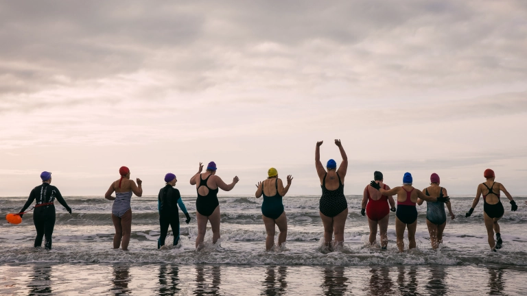 ten individuals going cold water swimming wearing swimsuits and swim caps. Seen from behind, they are stood in the shallow waves of the sea. One person is holding their hands above their head, others have their hands in the air.