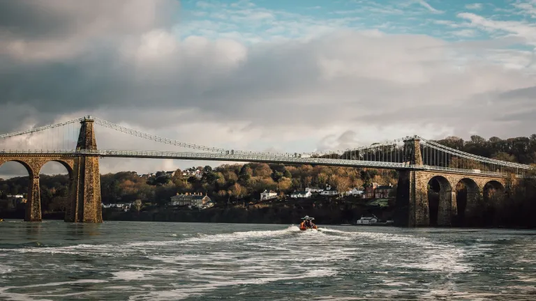 Suspension bridge with houses and trees in the distance, and a rib boat on the Menai Strait
