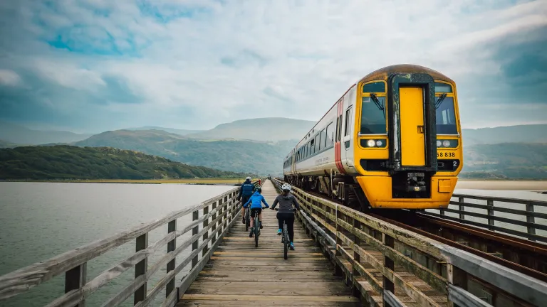 Three cyclists on a wooden railway bridge path, with a train passing by.