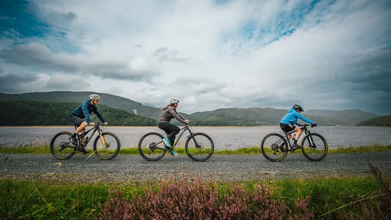 Three cyclists on a path by a river, with mountains in the background.