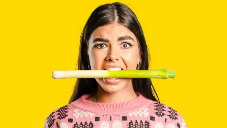 A woman wearing a pink jumper with a leek in her mouth.
