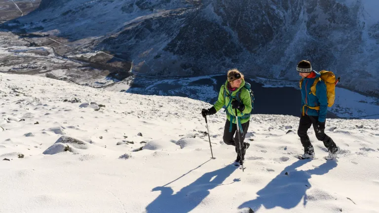 Two hikers walking on a snow-covered mountain summit with backpacks and poles, surrounded by rugged peaks under a partly cloudy sky.