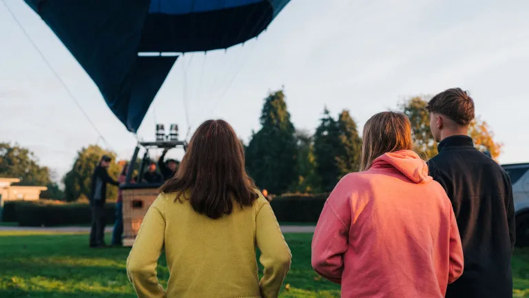 Three people standing on grass watching a hot air balloon being prepared for flight, with trees and a clear sky in the background.