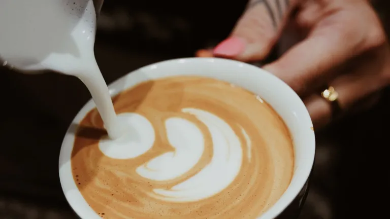 Here’s concise alt text for the second image — and as requested, no mention of any blurred face:  **Alt text:**   *A close-up of steamed milk being poured into a cup of coffee to create latte art and hand with tattoo and ring.