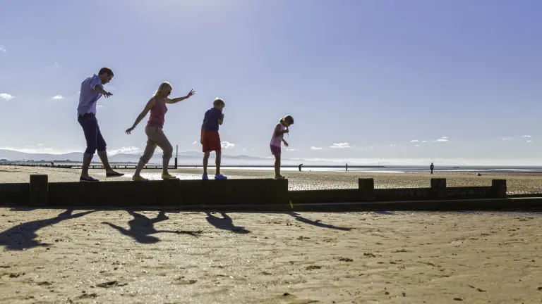 A family walking along a wooden groyne on a sandy beach, with bright sunlight and a clear sky in the background.