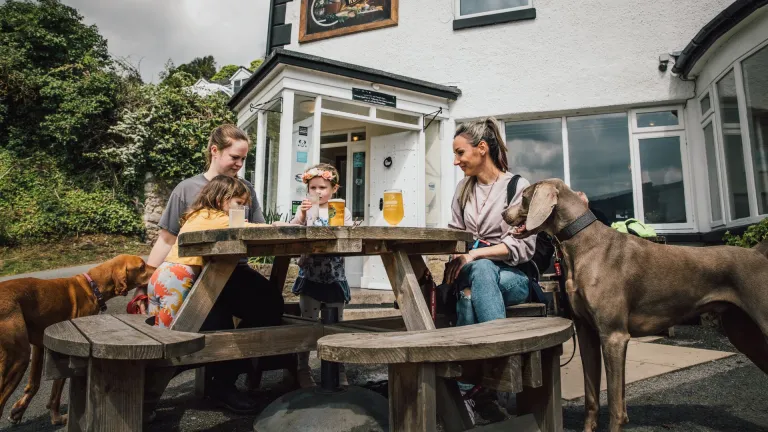 Family sit at a wooden picnic table outside a white pub with drinks, while two dogs stand nearby.