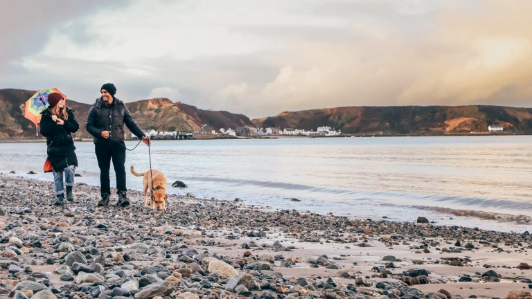 Couple and dog on a pebble beach beside a calm bay, with distant coastal village and hills under a cloudy sky.