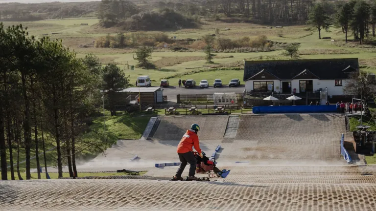 A child in an adapted sled / ski-pram being pushed by a man on a dry ski-slope.