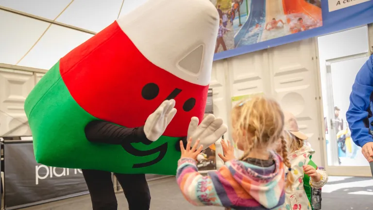 A costumed mascot in red and green leans down to greet a small child at an indoor event.