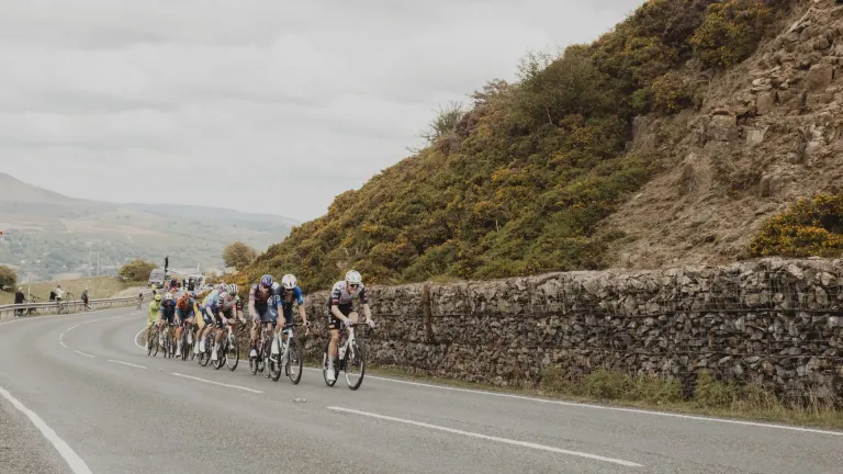 Professional cyclists racing in the 2025 Tour of Britain on the Tumble climb from Blaenavon, with a rocky hillside and gorse lining the road