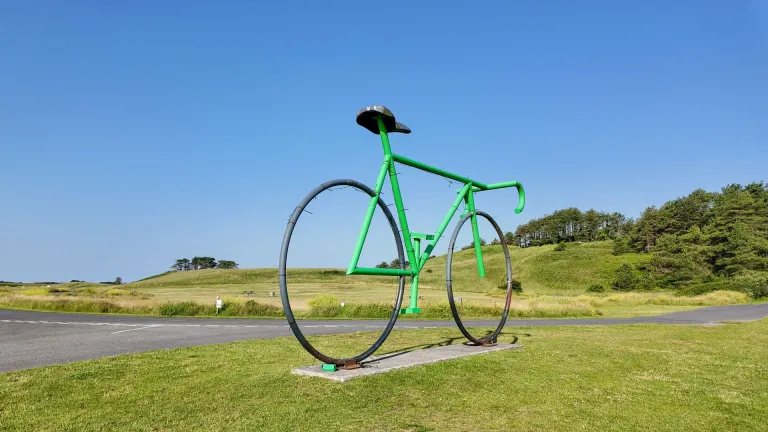 Green oversized bicycle sculpture standing beside parkland road in Pembrey Country Park.
