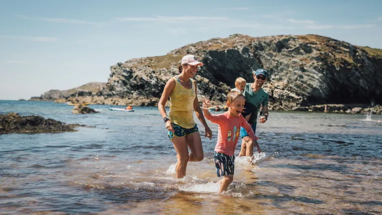 Family paddling and splashing in shallow water at a rocky cove on a sunny day, with kayaks visible offshore and rugged coastal cliffs in the background.
