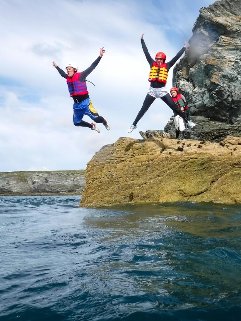 Two people wearing life jackets and helmets, coasteering and jumping from a rock into the sea