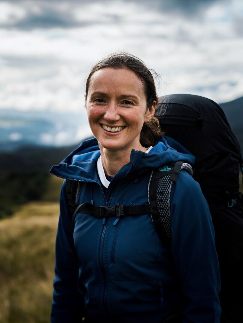Claire Copeman smiling at the camera wearing outdoor gear and carrying a backpack with mountains in the background.
