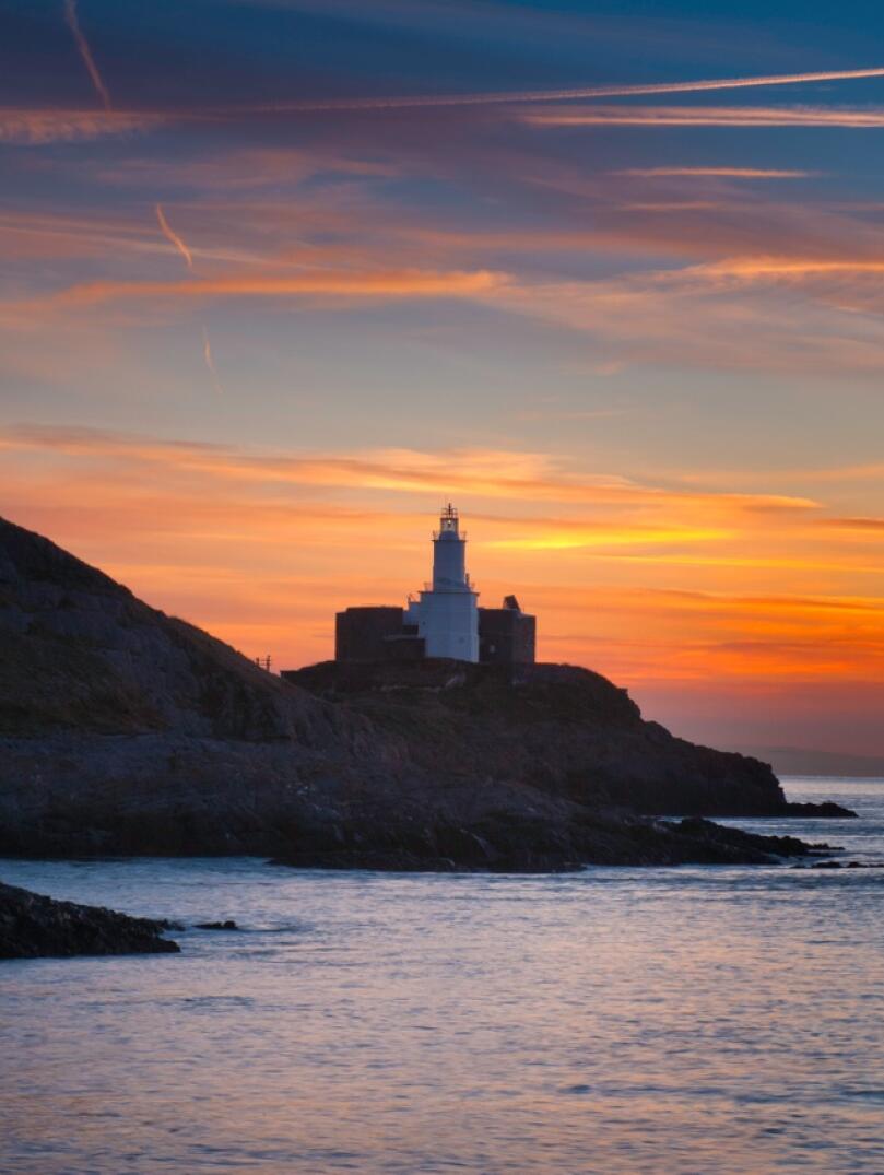 A lighthouse on the edge of a rocky headland against a backdrop of an orange sunrise.