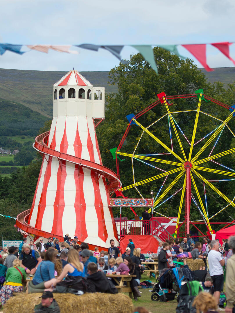 A red and white striped helter-skelter slide and a big wheel fairground ride in a field of festival goers.