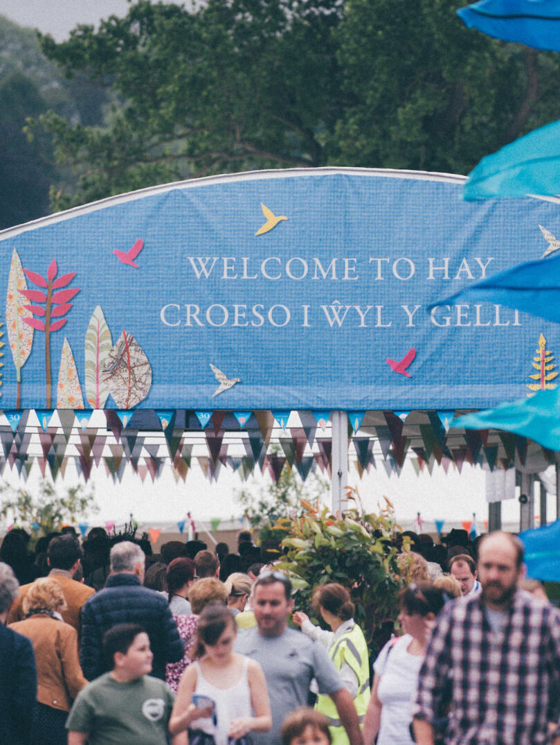 People walking  by The Hay Festival entrance with bunting flying in the breeze.