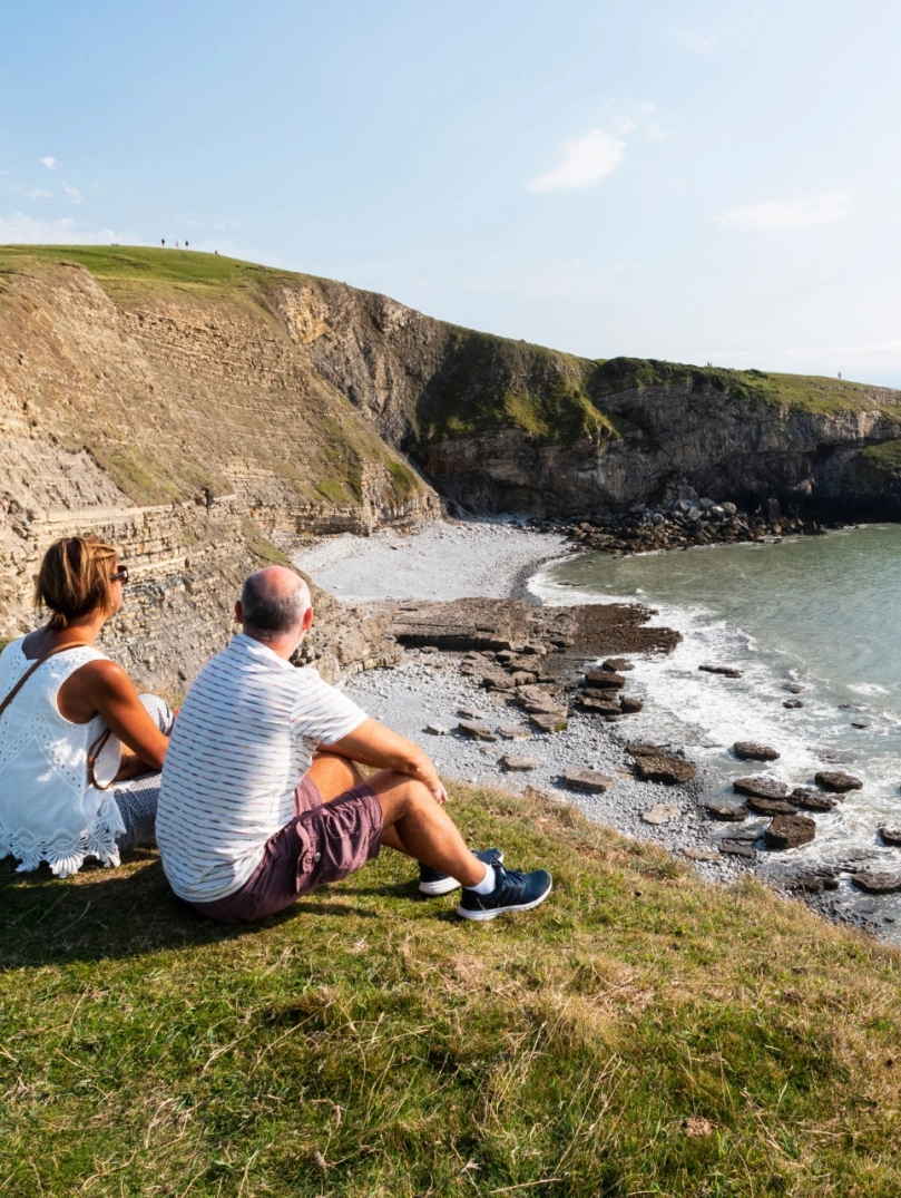 Man and woman sat on a clifftop looking out to sea.