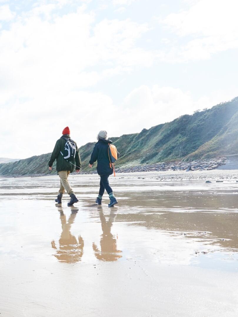 A man and a woman dressed warmly, walking on wet sand on a beach.