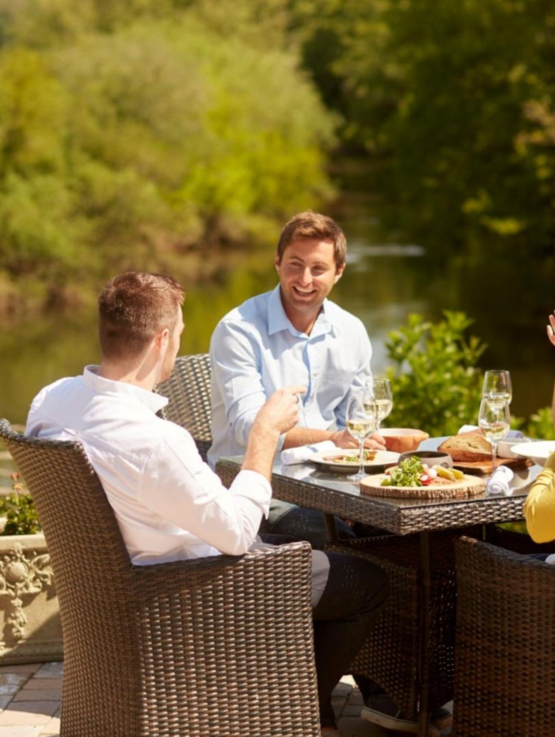 two men and two women in smart cauusal clothing sitting at a table with food and drink outside by the water with red flowers and green trees in the background. 