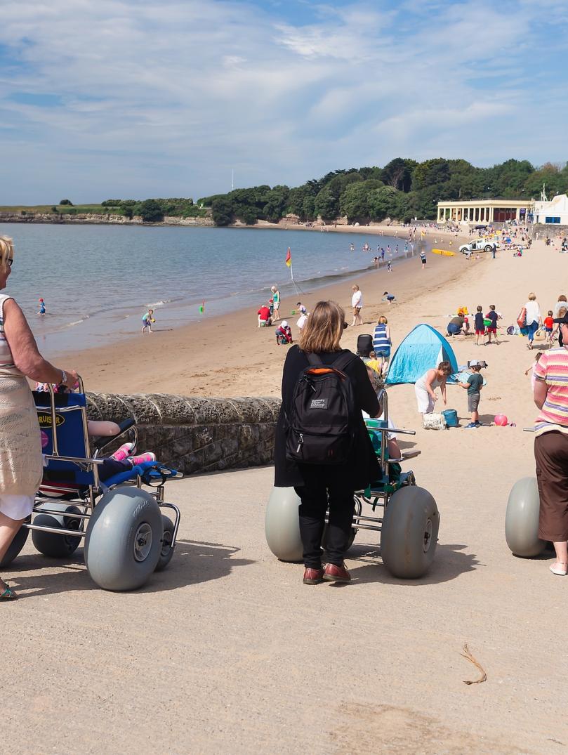 Three females pushing all-terrain beach wheelchairs down slope towards sandy beach.