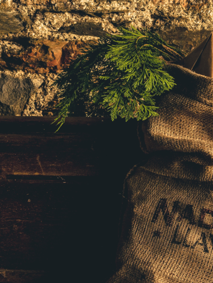 Brown hessian sack stuffed with christmas gifts and printed with the Welsh words Nadolig Llawen, hanging on a wooden mantle against a stone wall.