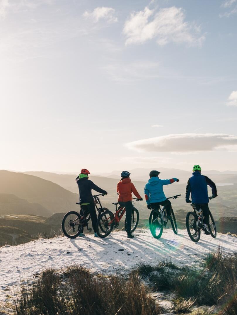 Four friends on mountain bikes on a snowy mountain top, looking out at the mountain scenery.