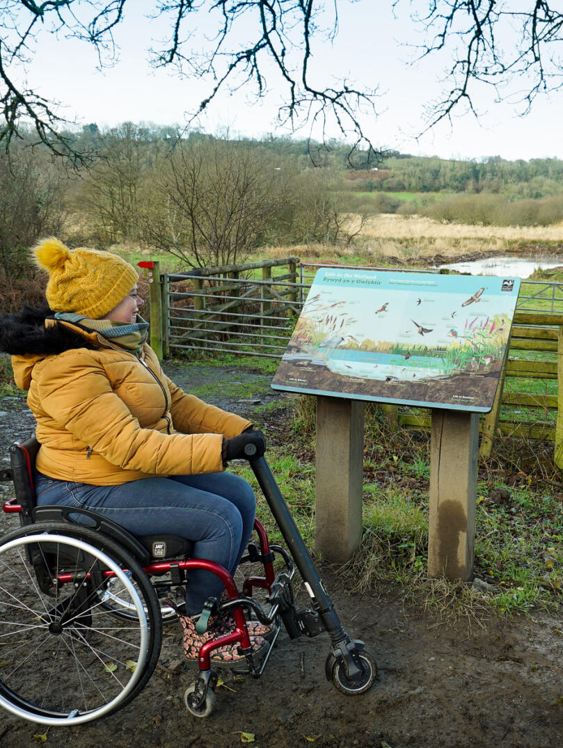 A woman using a wheelchair looking at an info board by a pond.