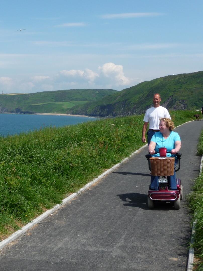 Lady on a mobility scooter with male companion on a tarmac section of the wales coast path looking out to sea