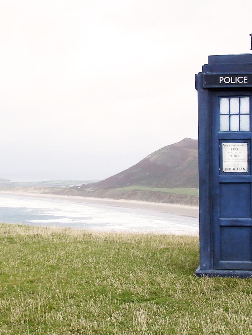 Blue police box film prop on grass with view of vast beach.