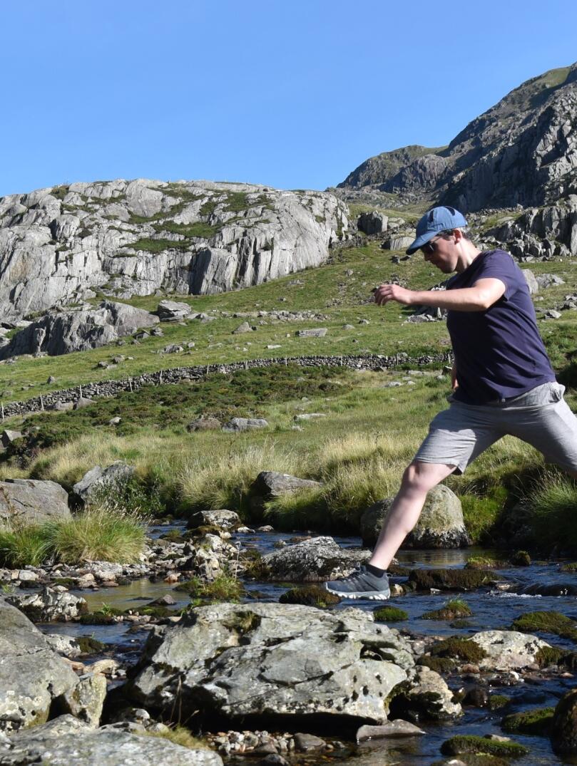 Mann springt über einen Fluss mit Steinen und blauem Himmel im Hintergrund.