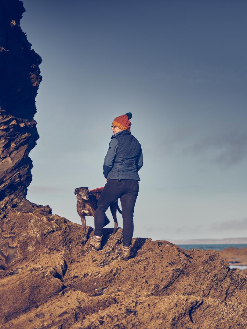 A person and a dog standing on rocks and looking out to sea.