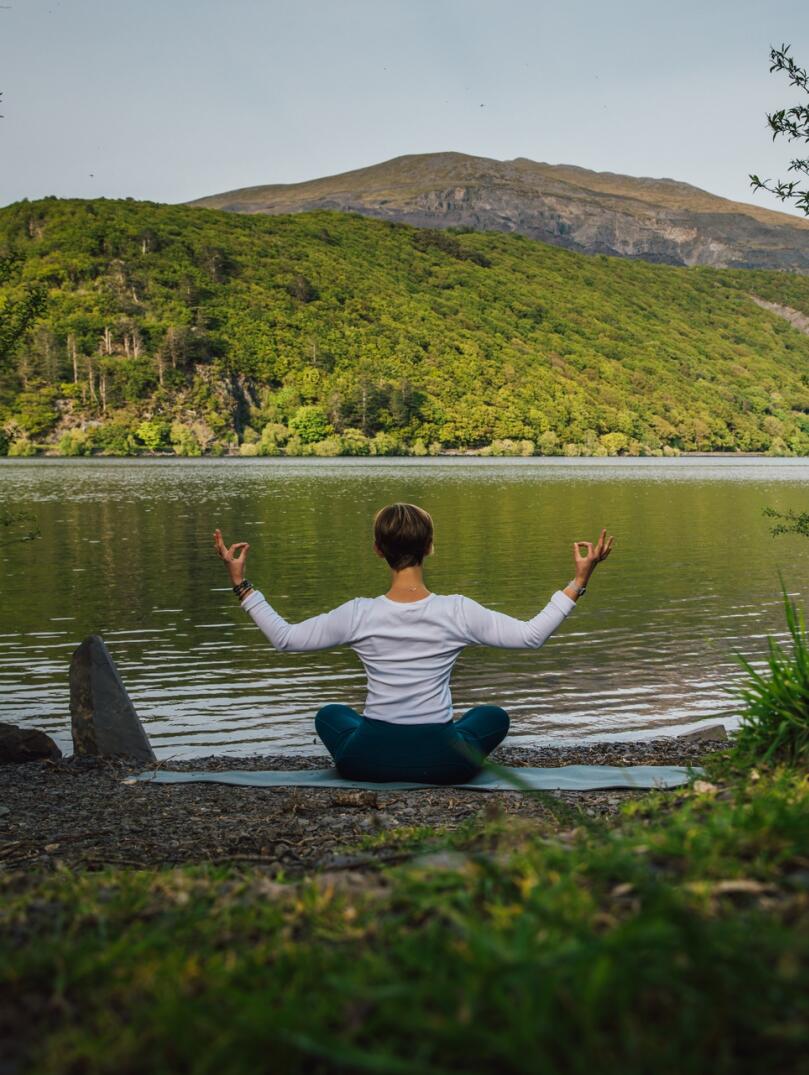 Person seen from behind, sat in a yoga pose in front of a lake with a mountain landscape in the distance