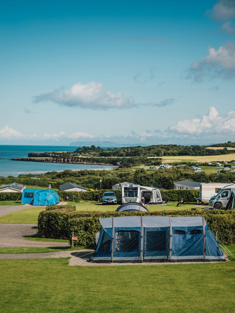 Campingplatz mit Zelten und Campervans auf einer Wiese mit Hecken und dem Meer im Hintergrund.